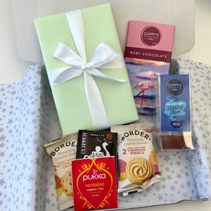 May include: A gift box filled with various items, including a green wrapped book, a chocolate bar with the text "GUPPY'S CHOCOLATES YORK RUBY CHOCOLATE", a box of biscuits with the text "BORDER. Beautifully Crafted Biscuits", a box of tea bags with the text "CLIPPER", a box of hot chocolate with the text "GUPPY'S CHOCOLATES YORK HOT CHOCOLATE Stirring Stick Milk", and a box of tea with the text "pukka revitalise organic/bio revital".
