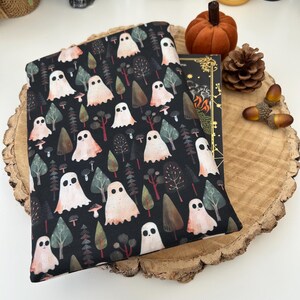 A black book sleeve with a repeating pattern of white ghosts, trees, and mushrooms. The sleeve is sat upon a book on top of a wooden slice. A pumpkin, pinecone, and acorns are in the background, creating a Halloween or autumn atmosphere.