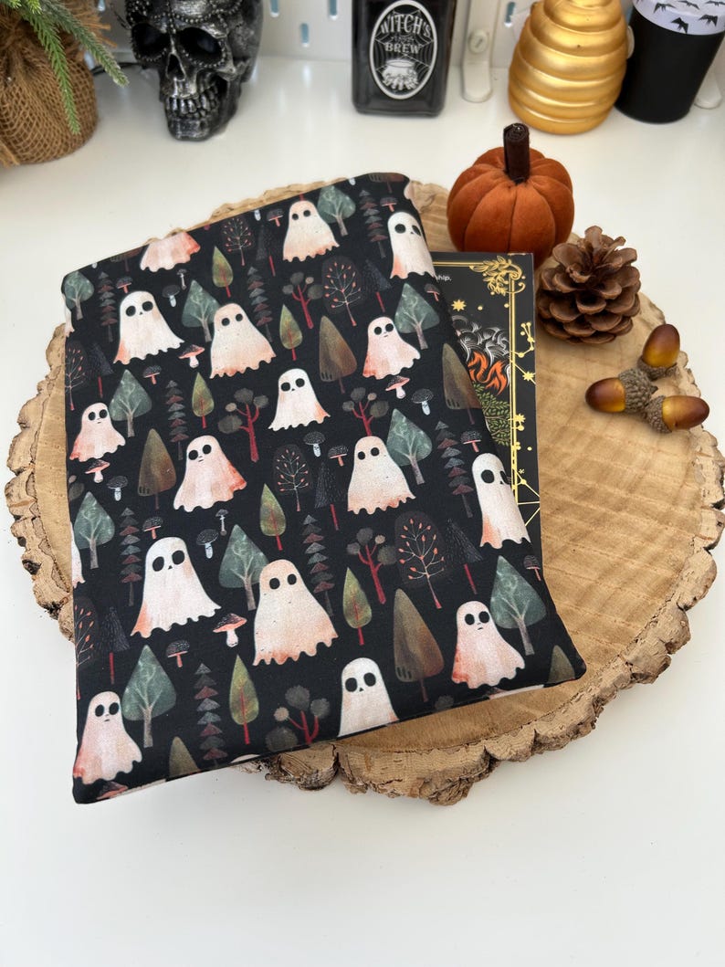 A black book sleeve with a repeating pattern of white ghosts, trees, and mushrooms. The sleeve is sat upon a book on top of a wooden slice. A pumpkin, pinecone, and acorns are in the background, creating a Halloween or autumn atmosphere.