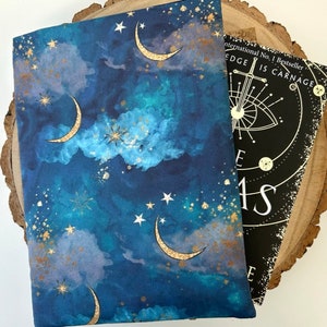 A dark blue book sleeve with a celestial design featuring gold  coloured crescent moons, stars, and clouds. The sleeve rests on a book. The book sleeve is made of fabric.