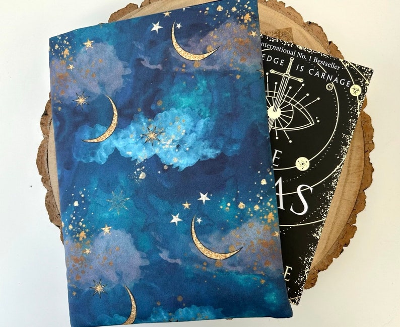 A dark blue book sleeve with a celestial design featuring gold  coloured crescent moons, stars, and clouds. The sleeve rests on a book. The book sleeve is made of fabric.