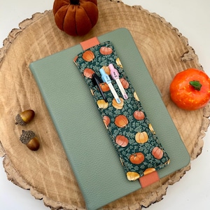 May include: A green notebook with a pumpkin-patterned pen holder, holding three pens. The pen holder features orange pumpkins on a dark green background. Two small pumpkins and acorns are also present.