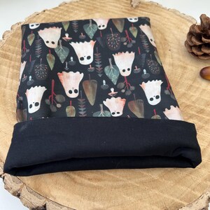 A black book sleeve with a turned-up edge to show the black lining. The book sleeve displays a repeating pattern of white ghost figures, green leaves, and brown acorns on a dark background. The item is shown on a wooden surface.