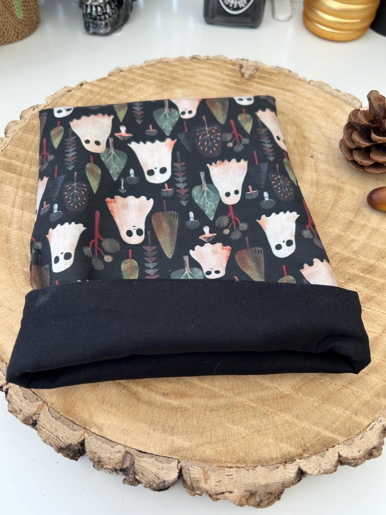 A black book sleeve with a turned-up edge to show the black lining. The book sleeve displays a repeating pattern of white ghost figures, green leaves, and brown acorns on a dark background. The item is shown on a wooden surface.