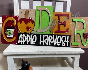 May include: A wooden sign with the word "CIDER" spelled out in colorful letters, with the words "APPLE HARVEST" below. The sign features apple and cinnamon stick illustrations, and is set on a small white chair with a black crow figurine.