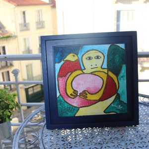 May include: A framed painting of a colorful abstract figure with a bird on its shoulder. The figure is yellow, red, and pink with a blue background. The painting is in a dark blue frame.