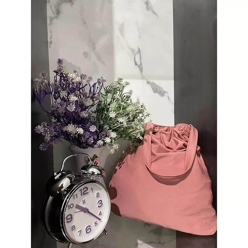 May include: A pink fabric tote bag with a drawstring closure and a silver alarm clock with a white face and black hands. The clock is set to 12:00. The bag and clock are on a white and gray marble surface.