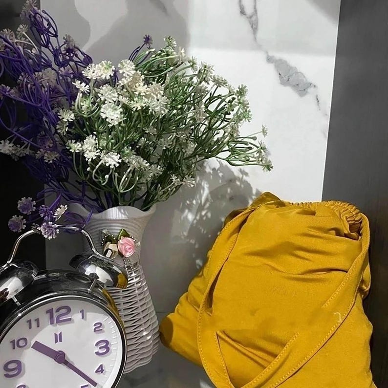 May include: A yellow fabric pouch with a drawstring closure. The pouch is sitting on a white surface next to a white vase with a pink flower and a silver alarm clock with a white face.