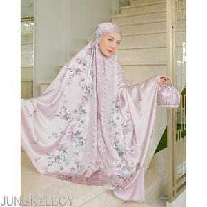 Pu&ograve; includere: Un completo da preghiera rosa chiaro con un motivo floreale, che include un copricapo, una veste e una piccola borsa abbinata. Il set presenta un design delicato con sfumature di rosa, viola e verde. L'indumento &egrave; indossato su una scala.