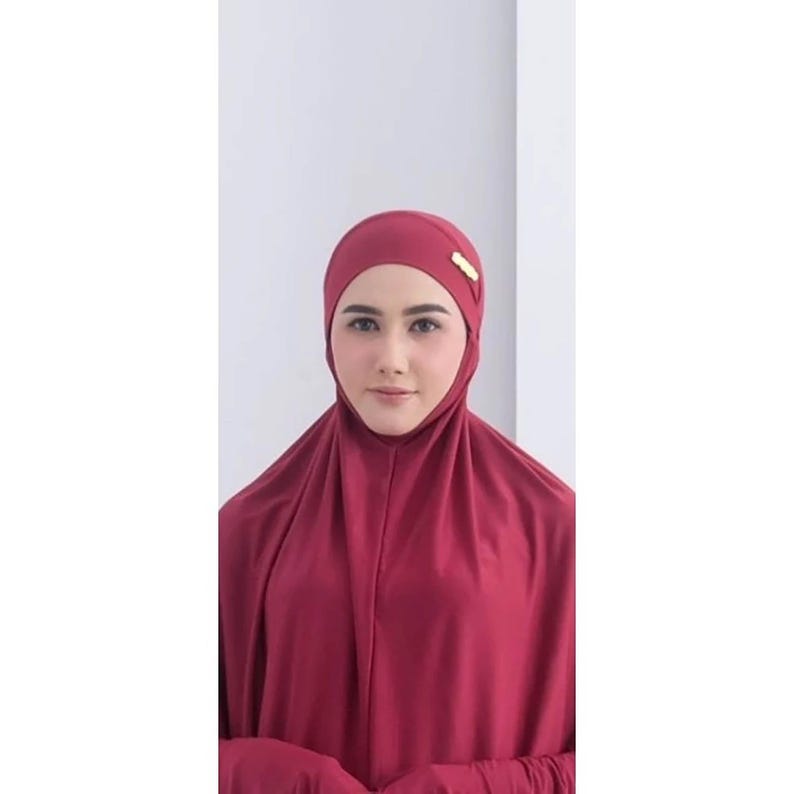 May include: A burgundy prayer dress with long sleeves and a matching head covering. The head covering has a gold accent.