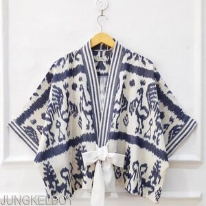 May include: A cream-colored kimono-style top with a blue patterned design featuring bird-like figures. The top has a white bow tie closure and striped accents on the lapel. The sleeves are wide and the fabric appears to be lightweight.