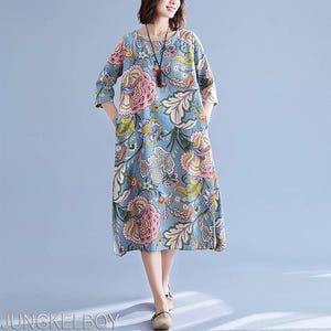 以下が含まれることがあります： ピンク、黄色、緑の花柄の、水色の膝丈ワンピース。 ワンピースは七分袖で、丸いネックラインです。 左下には「JUNGKELBOY」の文字があります。