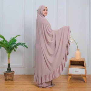 May include: A mauve prayer shawl with a ruffled hem. The shawl is draped over a person's shoulders and is worn with a matching mauve headscarf.