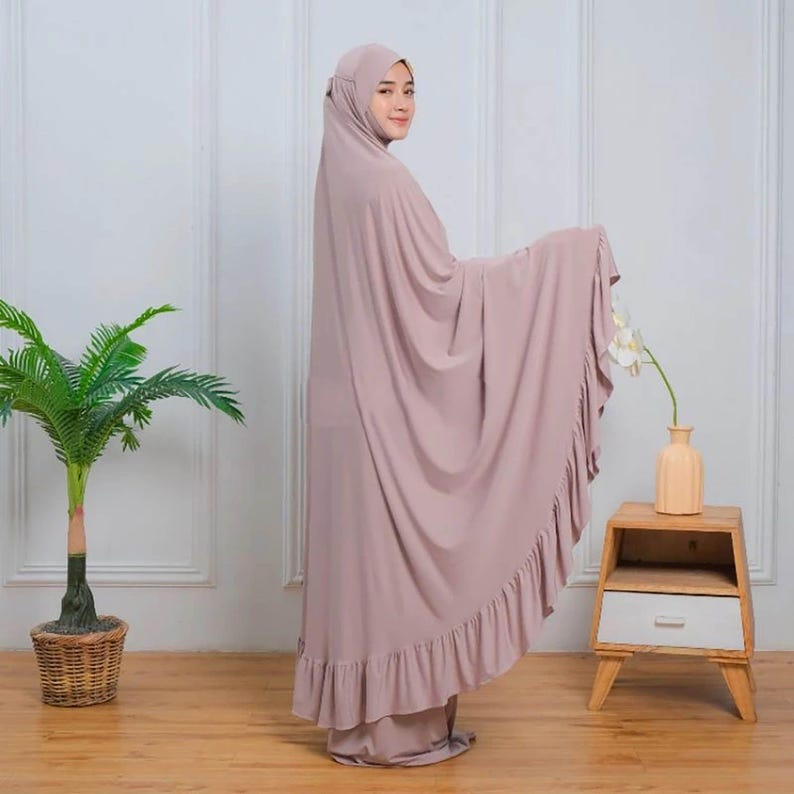 May include: A mauve prayer shawl with a ruffled hem. The shawl is draped over a person's shoulders and is worn with a matching mauve headscarf.