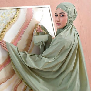 May include: A sage green prayer garment set, including a head covering and a flowing robe. The set is accessorized with a small matching purse with a gold chain. The background features an abstract painting with gold and pink accents.