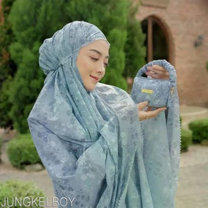 May include: A light blue floral patterned prayer garment and matching small handbag. The set includes a head covering and a long, flowing robe. The handbag has a short handle and a gold-coloured zipper. The text "JUNGKELBOY" is visible at the bottom.