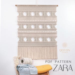 Puede incluir: Un tapiz de pared de macramé beige con patrones geométricos y flecos. El diseño incluye formas de diamante y bandas horizontales. El texto "PDF PATTERN ZARA" es visible. El tapiz se muestra en una pared blanca.