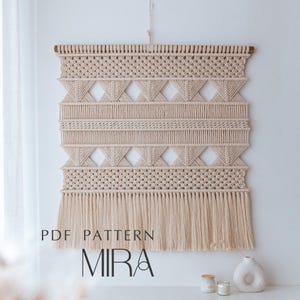 以下が含まれることがあります： 幾何学模様とフリンジが付いたベージュのマクラメ壁掛け。壁の装飾は、ダイヤモンドと水平線のデザインが特徴です。「PDF PATTERN MIRA」というテキストが画像の下部に表示されています。