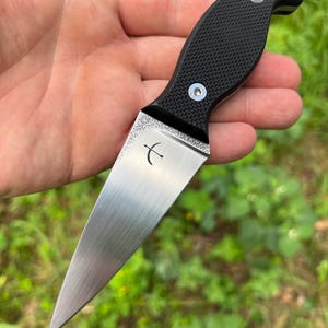 May include: A small, fixed-blade knife with a textured black handle and a silver blade. The blade has a curved edge and a small, etched design near the handle. The handle is secured with two silver rivets. The knife is designed for utility.