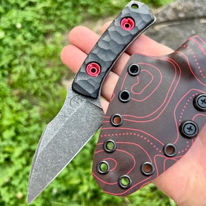 Puede incluir: Un cuchillo de hoja fija con mango gris oscuro texturizado, dos tornillos rojos brillantes y una hoja con acabado lavado a la piedra. Incluye una funda de Kydex marrón oscuro con un patrón de mapa topográfico rojo y ojales negros.