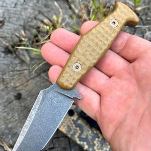 Könnte beinhalten: Ein kleines Messer mit einem tanfarbenen Griff mit strukturiertem Muster und einer grauen Klinge. Die Klinge hat eine Spitze und ein kleines, gebogenes Design. Das Messer wird in einer Hand vor einem unscharfen Hintergrund gehalten.