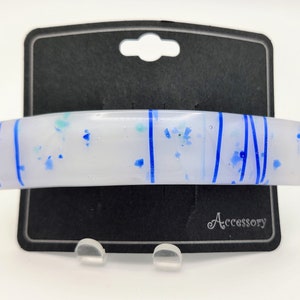 May include: White hair clip with blue and turquoise flecks and blue lines. The clip is on a black card with the word "Accessory" printed on it.