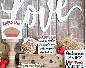 May include: A decorative fall-themed display featuring wooden signs with the words "Apple Pie," "Apple," and "Autumn Skies & Pies." A caramel apple and a large white "Love" sign are also present. The background is a rustic wooden wall.