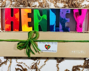 Puede incluir: Una caja marrón con una cinta verde atada alrededor. La caja contiene un conjunto de letras de crayón de colores que deletrean el nombre "HENLEY". La caja tiene una etiqueta que dice "Mimi hizo esto para ti" y una dirección de sitio web.