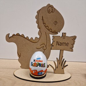 May include: A wooden dinosaur holding a sign that says "Name" and a Kinder Surprise egg. The dinosaur is brown and has a green grass base.