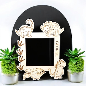 May include: A wooden picture frame featuring a dinosaur theme. The frame is decorated with four different dinosaurs, including a stegosaurus, a triceratops, a pterodactyl, and a tyrannosaurus rex. The frame has a space for a photo and the name "GEORGE" is written on the side.