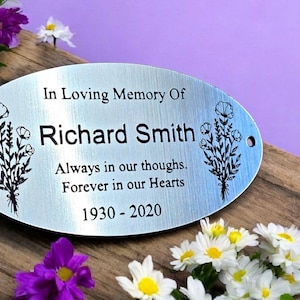 May include: A silver oval memorial plaque with engraved text: "In Loving Memory Of Richard Smith Always in our thoughts. Forever in our Hearts 1930-2020". The plaque features a floral design with two bouquets of flowers on either side of the text.