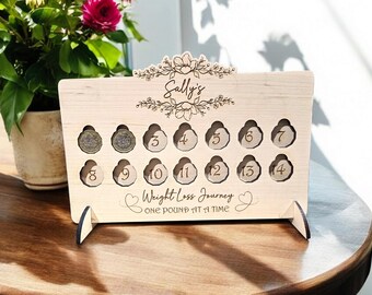 May include: A wooden weight loss tracker with a floral design and the name "Sally's" engraved. It has 14 slots for coins, numbered 3 through 14, and the text "Weight Loss Journey One Pound At A Time".