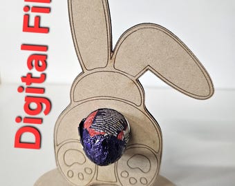 May include: A wooden bunny rabbit cutout with a chocolate egg in the center. The bunny has a round base with paw prints. The bunny is brown and the egg is purple and red.