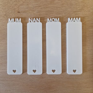 May include: Four white rectangular bookmarks with heart-shaped cutouts at the bottom. Each bookmark has a different word at the top: MUM, NAN, MOM, and MAM. The bookmarks are on a light brown wooden surface.