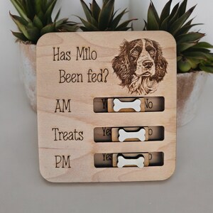 May include: Wooden dog feeding tracker with a picture of a brown and white dog. The tracker has a sliding bone to indicate if the dog has been fed in the AM, PM, or for treats.