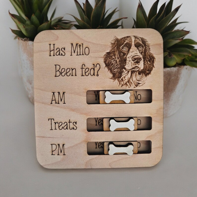 May include: Wooden dog feeding tracker with a picture of a brown and white dog. The tracker has a sliding bone to indicate if the dog has been fed in the AM, PM, or for treats.