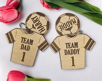 Puede incluir: Dos llaveros de madera con forma de camisetas de fútbol con las palabras "Team Dad 1" y "Team Daddy 1" en ellos. Cada llavero tiene un charm de balón de fútbol de madera con los nombres "Harry" y "Maddie" en ellos.