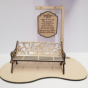 May include: A wooden Christmas in Heaven bench scene. The bench has an ornate design and is positioned in front of a sign that reads "Christmas in Heaven." The sign includes a poem about loved ones. The scene is made of light-colored wood.