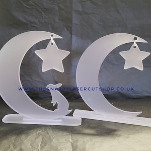 Peut inclure: Deux décorations en acrylique blanc en forme de croissant de lune et d'étoile avec une silhouette de lapin sur l'une d'elles. Les décorations ont un petit trou en haut pour être suspendues. Le texte "www.thesnappyshop.co.uk" est visible en bas de l'image.