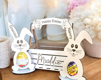 May include: Wooden Easter bunny egg holders with personalized names "Grandad", "Maddie", and "Nanny". The bunnies are holding chocolate eggs and there is a banner that says "Happy Easter".