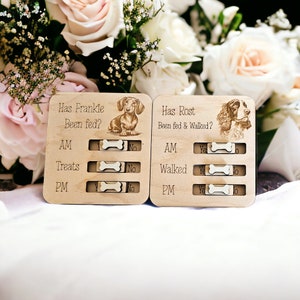 May include: Two wooden dog care trackers with sliding toggles. The left tracker is for Frankie and asks "Has Frankie Been Fed?" with options for AM, Treats, and PM. The right tracker is for Rost and asks "Has Rost Been Fed & Walked?" with options for AM, Walked, and PM.