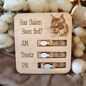 May include: Wooden sign with a cat illustration and the text "Has Salem Been fed?" with a sliding toggle for AM, Treats, and PM. Each toggle has a fish icon and a "No" label.
