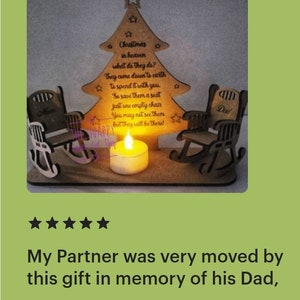 Puede incluir: Un adorno de &aacute;rbol de Navidad de madera con dos mecedoras, una etiquetada como "Mam&aacute;" y la otra como "Pap&aacute;". El &aacute;rbol tiene una cita sobre los seres queridos en el cielo y una vela encendida frente a &eacute;l.