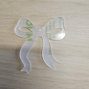 May include: A clear, bow-shaped acrylic cutout with a ribbon design. The bow has a small hole in the centre and is resting on a light wood surface. The acrylic has a protective film with green and white text.