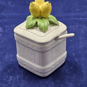 May include: A white ceramic sugar bowl with a lid, featuring a lemon-shaped handle. The bowl has a square shape with a wooden texture design. A small spoon rests on the side.