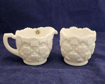 May include: A white milk glass creamer and sugar bowl set. The pieces have a textured, geometric pattern and scalloped edges. The creamer has a handle and spout. The sugar bowl is open. A small sticker is visible on the creamer.