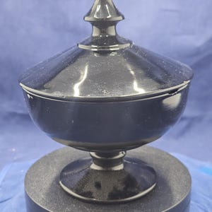 May include: A glossy black glass candy dish with a matching lid featuring a pointed finial. The bowl has a rounded shape and sits on a circular pedestal base. The lid is wide and sloped, fitting securely over the top of the bowl.