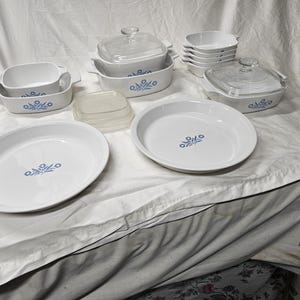 May include: A collection of white ceramic cookware with blue floral designs. The set includes various casserole dishes with lids, oval and round baking dishes, and small square dishes. The dishes are arranged on a white surface.