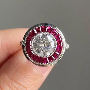 Puede incluir: Un anillo de plata con un gran diamante redondo central rodeado por un halo de rubíes rojos.