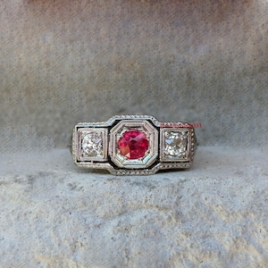 May include: A silver ring with a red gemstone and two smaller white gemstones. The ring has a detailed design with intricate carvings.